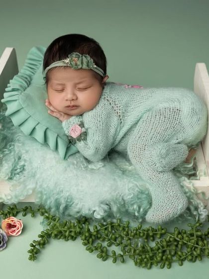A baby girl in a mint green knit outfit sleeps peacefully on her side in a miniature white bed prop. The greenery and flowers around the bed add a touch of nature.