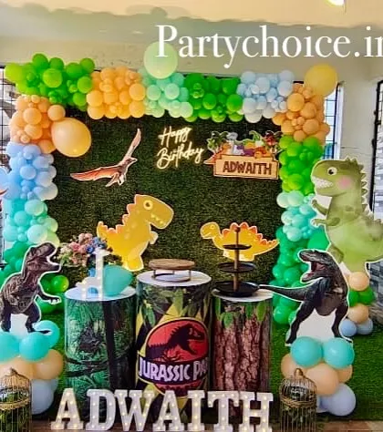 A dinosaur-themed birthday party setup in an outdoor covered area. It features a green wall backdrop, various dinosaur cutouts, and Jurassic Park-themed plinths.