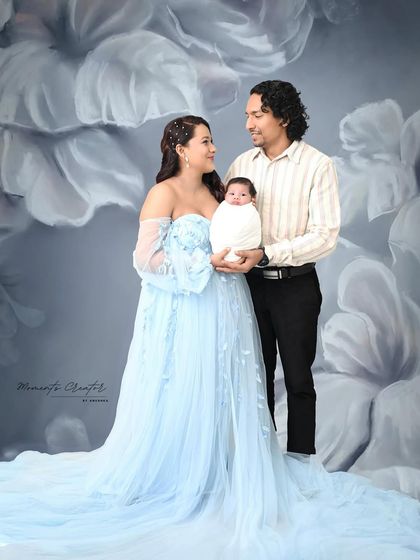 A classic family portrait with a newborn. The parents stand together holding their baby, dressed in a beautiful blue gown that matches the artistic floral backdrop.
