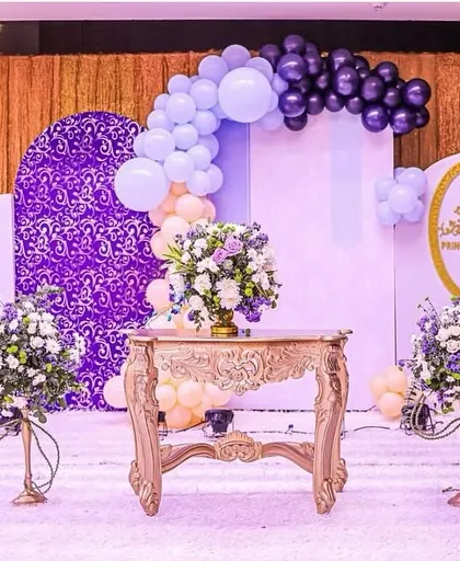 A full view of the royal prince stage setup. The design features multiple backdrops in purple and white, an ornate golden table, and elegant floral stands, all tied together with balloon arches.