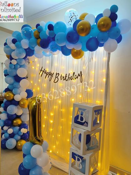A first birthday setup at home, featuring a blue and gold balloon arch, 'BABY' blocks, and a number '1' foil balloon.