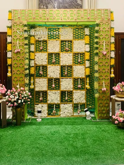 A modern and stylish backdrop featuring a green and gold jaali frame. The center is a checkerboard of white flowers, with hanging lotus buds adding a delicate, traditional touch.