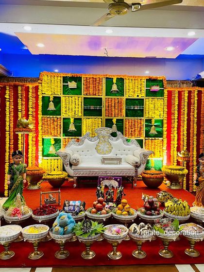 A vibrant and colorful stage decoration for a baby shower or half-saree function. The backdrop features a traditional checkered pattern with temple bells, all in bright festive colors.