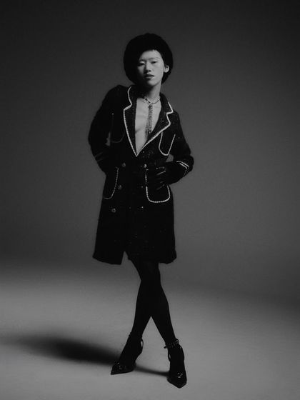 A black and white editorial shot featuring a Chanel-inspired look. The styling is classic and luxurious, with a focus on the tailored coat, fur details, and dramatic silhouette.