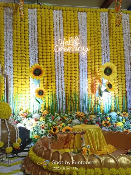 A popular and modern Haldi decor theme. This setup features a full wall of yellow and white marigold strings, decorated with sunflowers and a 'Haldi Ceremony' neon sign.