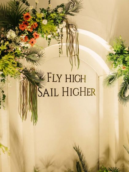 This custom arched backdrop for a joint 50th and 80th birthday features a personal quote, "Fly High Sail Higher," framed by lush tropical greenery and florals, making it a meaningful centerpiece.