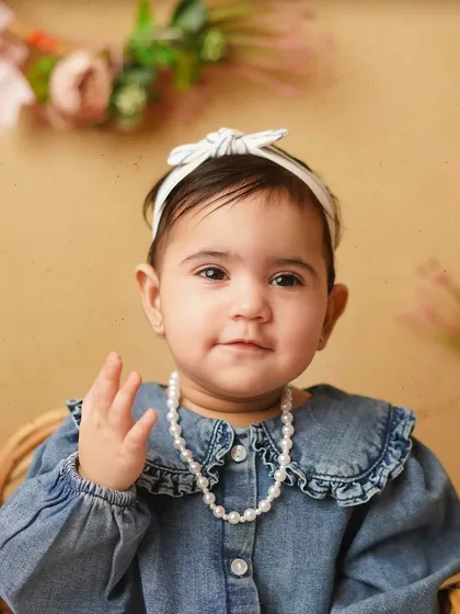 A close-up of a sweet baby girl, dressed in a denim shirt and pearls, giving a little wave. Her personality shines in this adorable portrait.