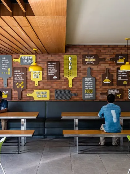 The cafeteria at the Stainless Steel Office features a vibrant and casual design. A brick wall is adorned with custom graphics, and the space offers a mix of bench seating and chairs in a bright, welcoming environment.