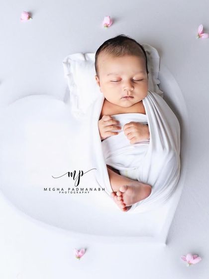 A baby sleeps in a white heart-shaped prop, surrounded by delicate pink petals. A beautiful and serene image.