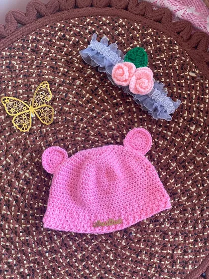 This image shows the pink bear-ear beanie next to a delicate crochet flower headband. Both are handmade and make a beautiful gift set for a baby.