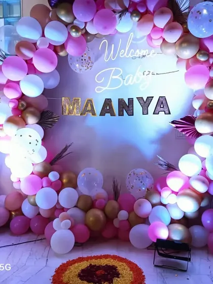 A vibrant welcome decoration for baby Maanya, featuring a full ring of pink, white, and gold balloons. A large teddy bear and personalized 'BABY' blocks make this a perfect photo spot.
