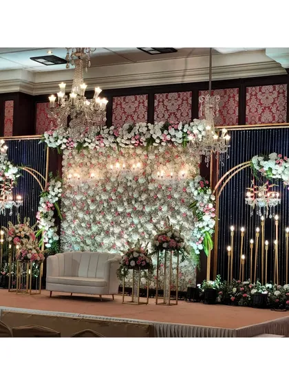 This elegant reception stage features a full wall of white and blush florals, framed by modern gold arches and accented with hanging crystal chandeliers. The design combines classic romance with contemporary style, providing a luxurious backdrop for the couple.