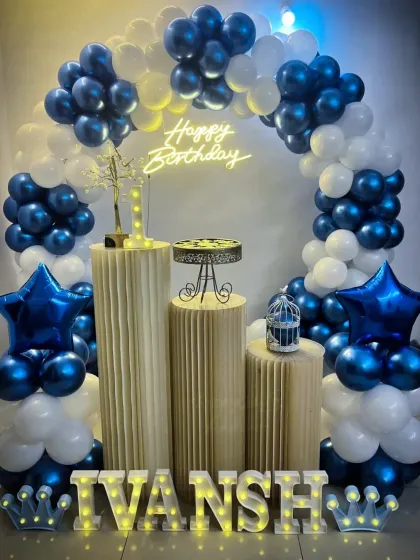 A stylish birthday party with a blue and white balloon arch, plinths, and star-shaped foil balloons. The name 'Ivansh' is spelled out in marquee letters.