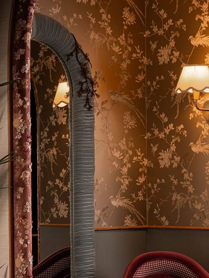 The reflection in the pleated mirror shows the layers of design in the boutique. The floral wallpaper, fabric-wrapped arch, and warm sconce lighting all contribute to the soft, boudoir-inspired mood of this intimate corner.