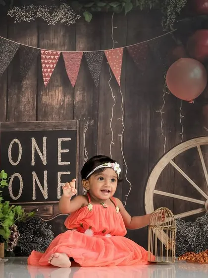 A rustic-themed first birthday shoot, with the baby girl in a coral dress against a wooden backdrop with a wagon wheel.
