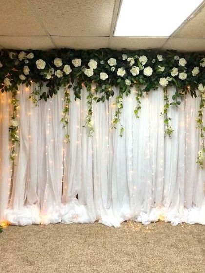 A simple and elegant flower decoration for a baptism. The backdrop consists of white drapes with a garland of white roses and greenery, lit by fairy lights.