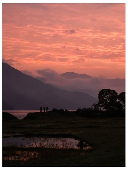 A stunning pink and purple sunset over the hills surrounding Phewa Lake, with silhouettes of people enjoying the view.