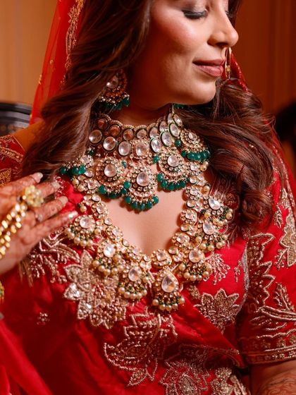 A close up shot focusing on the bride's magnificent jewelry. The layered Kundan necklace with green stones perfectly complements her red embroidered lehenga, showcasing royal elegance.