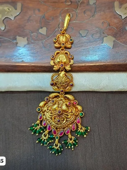 A beautiful 92.5 silver, gold-plated maang tikka featuring a traditional peacock design. The piece is embellished with red and green stones and green bead drops, perfect for a festive look.