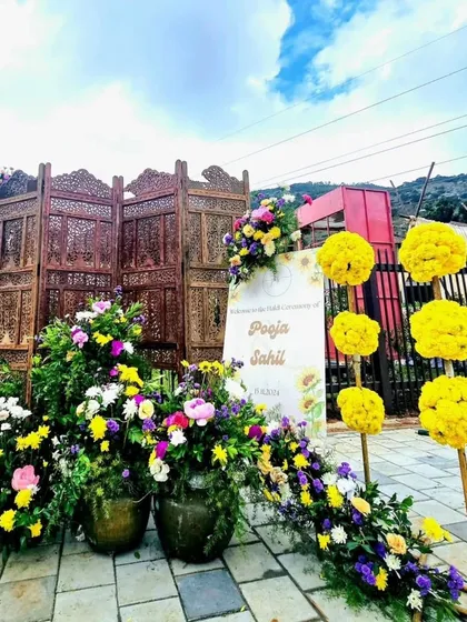 A simple and rustic welcome sign for a pre-wedding event. The sign is surrounded by colorful flowers and set against a carved wooden screen, creating a warm and inviting entrance.