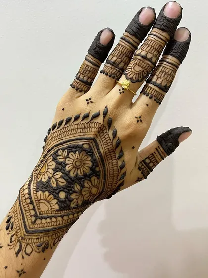 A bold and dark henna design featuring a large, filled floral pattern. The fingers are heavily detailed, creating a very rich and traditional look.
