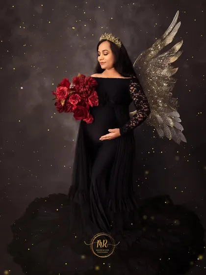 A dark and dreamy fine art portrait. The expectant mother wears a black gown and a crown, holding a bouquet of red roses, with golden angel wings adding a touch of magic.