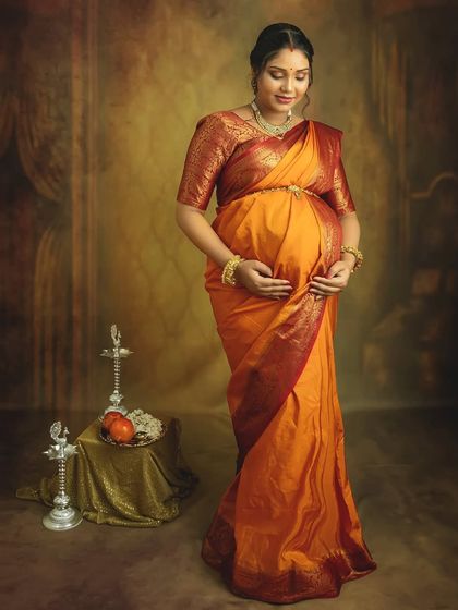 A serene solo portrait in a traditional orange saree. Cradling your bump, you look absolutely radiant against the classic studio backdrop with cultural props.