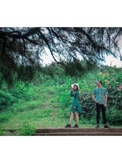 A couple stands on a stone path in a forest, framed by overhanging branches, creating a beautiful, nature-filled pre-wedding portrait.