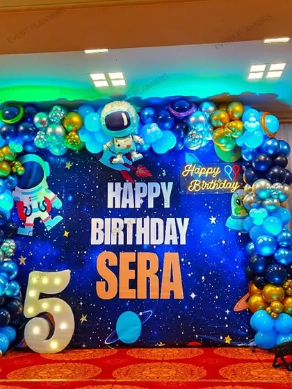 Blast off with an out-of-this-world space and astronaut themed birthday party. This large-scale setup features a galaxy backdrop, planets, astronaut cutouts, and a blue and gold balloon garland.