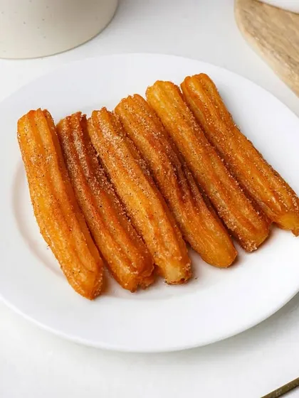 A plate of freshly made churros dusted with cinnamon sugar, with a side of chocolate dipping sauce. The clean white plate makes the golden-brown churros look extra crispy.