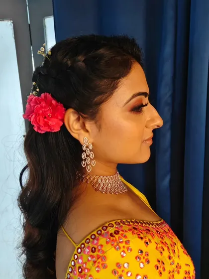 This half-up, half-down hairstyle for a Haldi ceremony is both beautiful and practical. It features a braided crown and soft waves, decorated with baby's breath and a single bright flower for a pop of color.