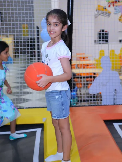 A young girl takes a break from bouncing to practice her basketball skills in the trampoline park. It's a great way to combine sports and play.
