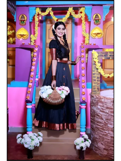 A beautiful shot of a model in a traditional black and gold half-saree, posing in a colorful, village-themed studio set. This look is perfect for celebrating South Indian heritage.