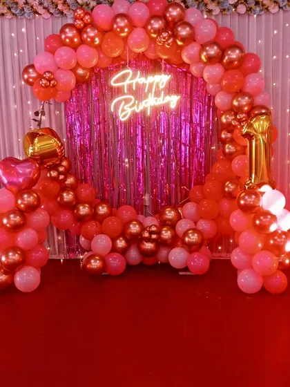 A vibrant pink and rose gold first birthday setup. This decoration features a balloon arch over a shimmery pink backdrop, a "Happy Birthday" neon sign, and a number "1" foil balloon.