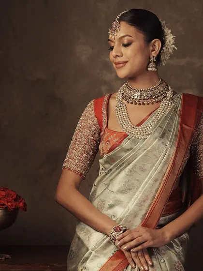 A serene portrait of a model in a light-colored saree with a red border. Her red blouse features intricate, all-over embroidery on the sleeves for a balanced and elegant look.