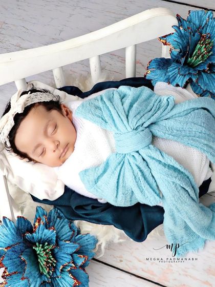 A baby wrapped in a blue bow, resting on a white diwan with dramatic blue flowers.