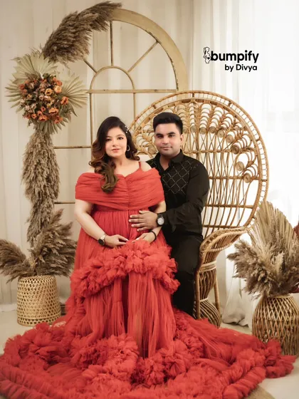 The couple together in the red gown setup. His hand rests gently on her bump, a simple gesture full of love and excitement for their journey ahead.