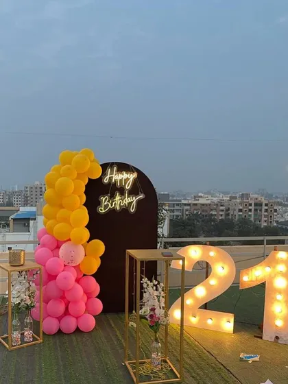 This is a duplicate of ID 21, 60, 67, 91, 108, 121, 168, 206, 232. A vibrant 21st birthday decoration on a rooftop. This setup features a balloon arrangement in hot pink and yellow, a 'Happy Birthday' neon sign, and large marquee numbers against a city skyline.