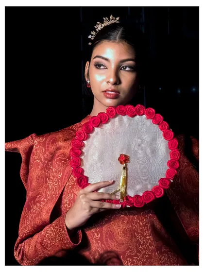 A close-up portrait of a model holding a beautifully crafted fan. This image highlights the accessories and the intricate details of the red brocade outfit, capturing a moment of elegance on the ramp.
