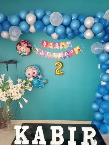 A simple Cocomelon decoration for a home setting. A blue and white balloon garland frames a 'Happy Birthday' banner and character foil balloons.