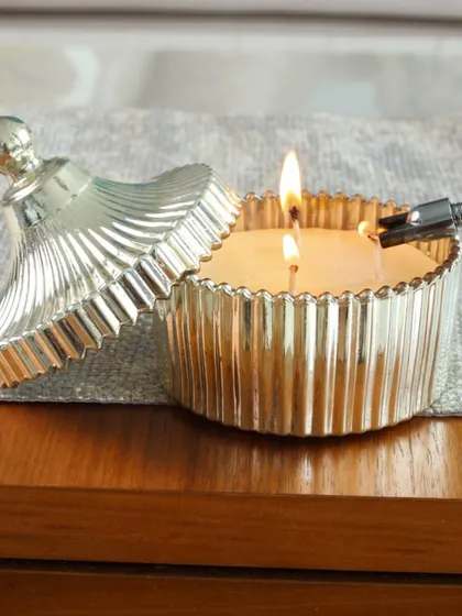 Our silver ribbed candle holder with a matching lid, its two wicks lit. The metallic finish and textured design create a warm, shimmering glow that enhances any atmosphere.