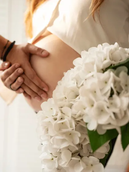 A close-up shot focusing on the hands of the couple gently holding the baby bump, with a bouquet of white flowers adding a touch of elegance and softness.