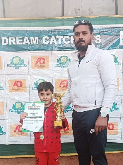 A proud coach with one of our youngest champions in the U6 category. The player holds his winner's trophy and certificate in front of the Dream Catcher's tournament banner.