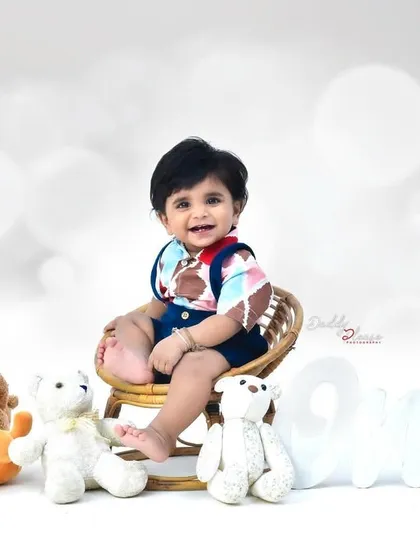 A happy baby boy sits on a small wicker chair, surrounded by a collection of cute teddy bears. The clean, white background keeps the focus on his adorable smile.