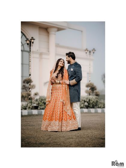 A full-length portrait of the couple in a beautifully manicured garden. Their traditional orange and navy blue outfits create a lovely contrast against the green and white background.