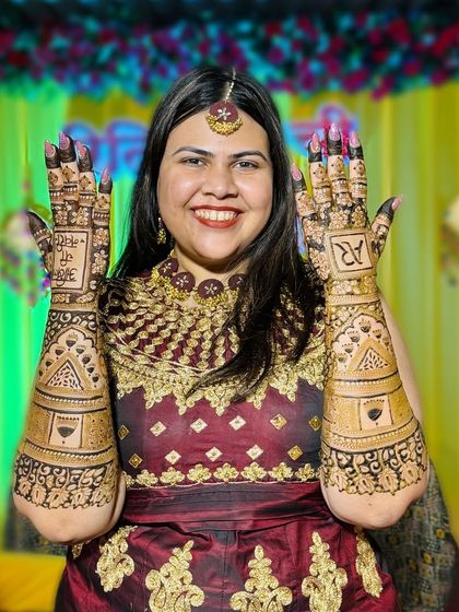 A beautiful bride proudly displaying her full-arm bridal mehendi. The design includes her and her partner's initials, 'AR', and a special message.