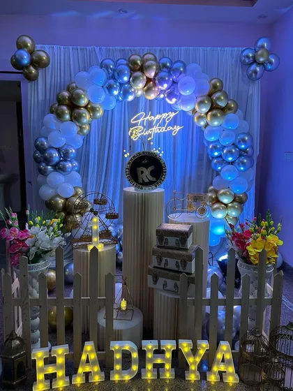 An elegant indoor setup with a gold and silver balloon arch. The decor includes a neon sign, stylish plinths, and marquee letters.