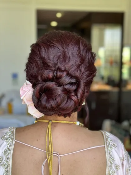 A detailed shot of a bridal bun with intricate twists and braids, colored in a rich burgundy. A single pink rose adds a soft, romantic touch to this elegant updo.