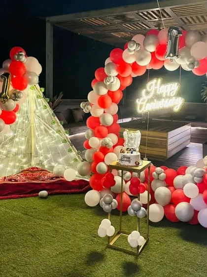A first-anniversary party on a terrace with an artificial lawn. The setup features a circular balloon arch in red and white, a neon sign, and a cozy, lit-up teepee tent.