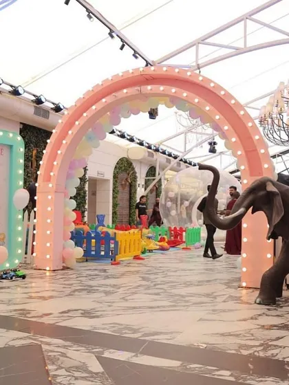 A grand entrance to a pastel carnival party, featuring a large rainbow arch, a light-up welcome sign, and a colorful play area, transforming an indoor venue into a magical space.